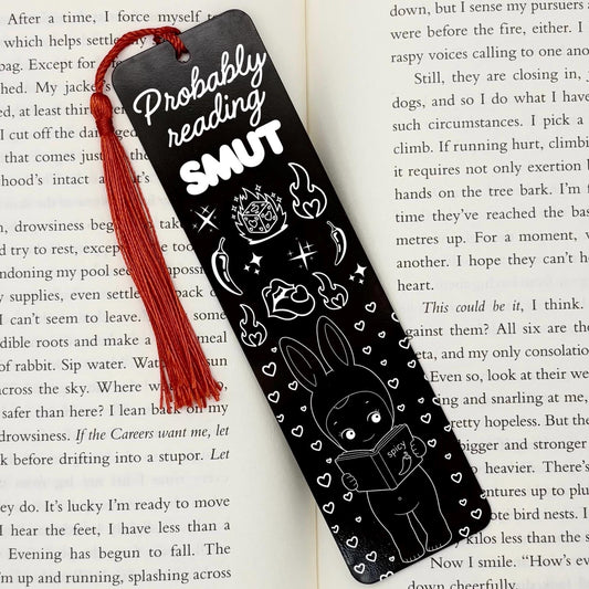 A black Probably Reading Smut Metal Bookmark on an open book with red tassel