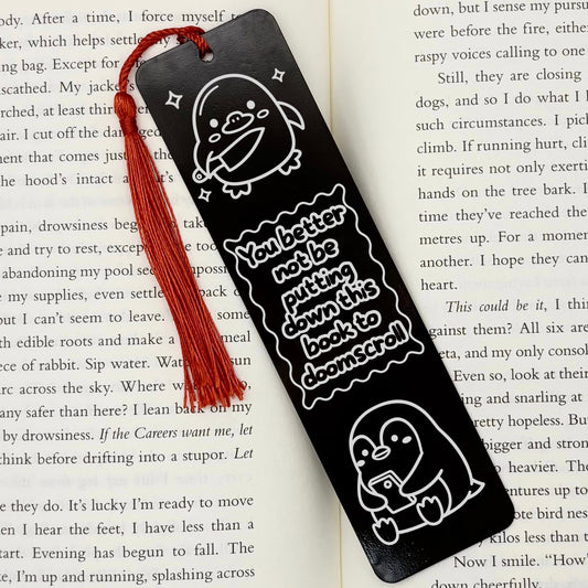 A black Doomscrolling Duck Metal Bookmark with red tassel on open book