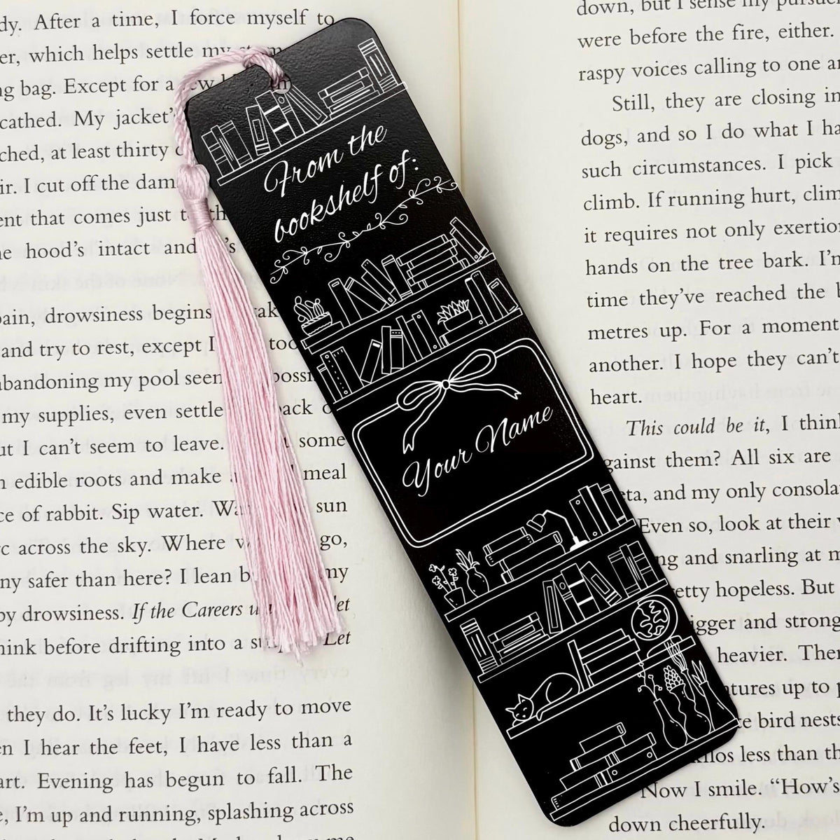 A black Personalised Name Bookshelf Metal Bookmark on an open book with a pink tassel