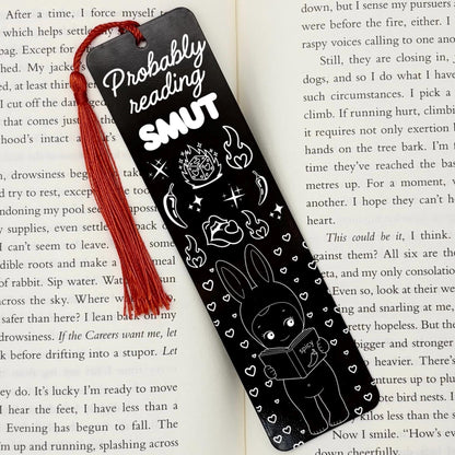A black Probably Reading Smut Metal Bookmark on an open book with red tassel