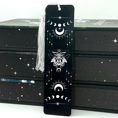 A black Celestial Moon and Stars Metal Bookmark with white tassel standing against a backdrop of books