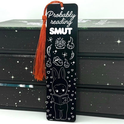 A black Probably Reading Smut Metal Bookmark with red tassel standing against a stack of books