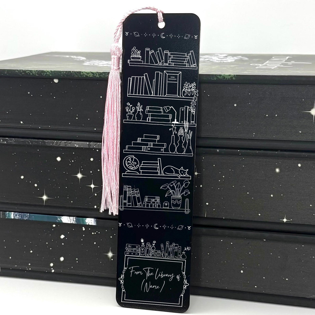 A black Personalised Name Library Shelf Metal Bookmark with pink tassel standing against a backdrop of books
