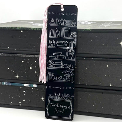 A black Personalised Name Library Shelf Metal Bookmark with pink tassel standing against a backdrop of books