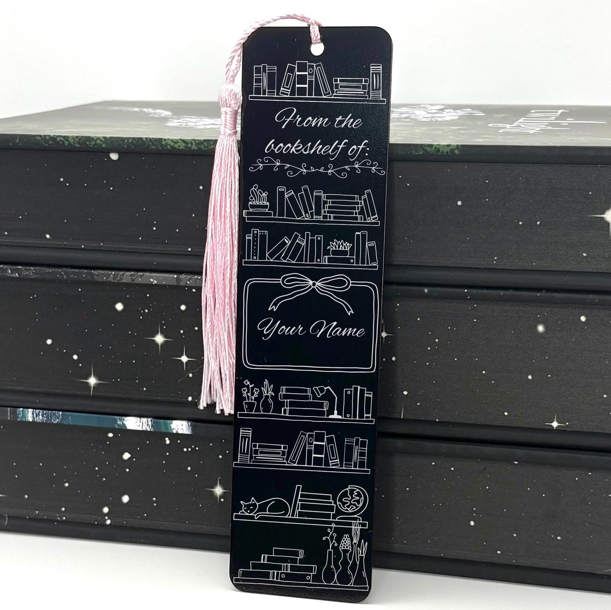 A black Personalised Name Bookshelf Metal Bookmark with pink tassel standing against a backdrop of books