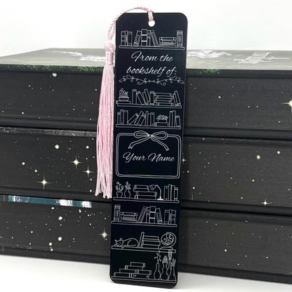 A black Personalised Name Bookshelf Metal Bookmark with pink tassel standing against a backdrop of books