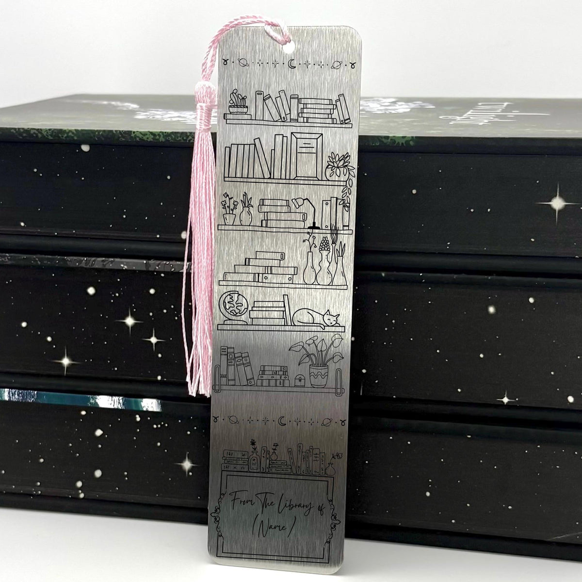 A brushed silver Personalised Name Library Shelf Metal Bookmark with pink tassel standing against a backdrop of books