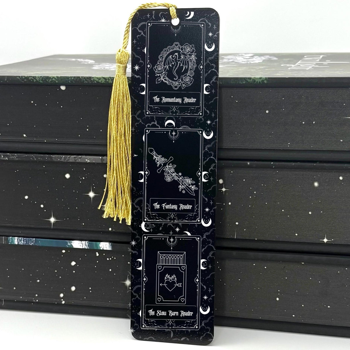 A black Fantasy Romantasy Slow Burn Metal Bookmark with gold tassel standing against a backdrop of books