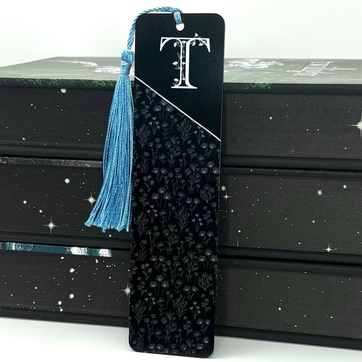 A black Personalised Floral Initial Metal Bookmark with light blue tassel standing against stack of books