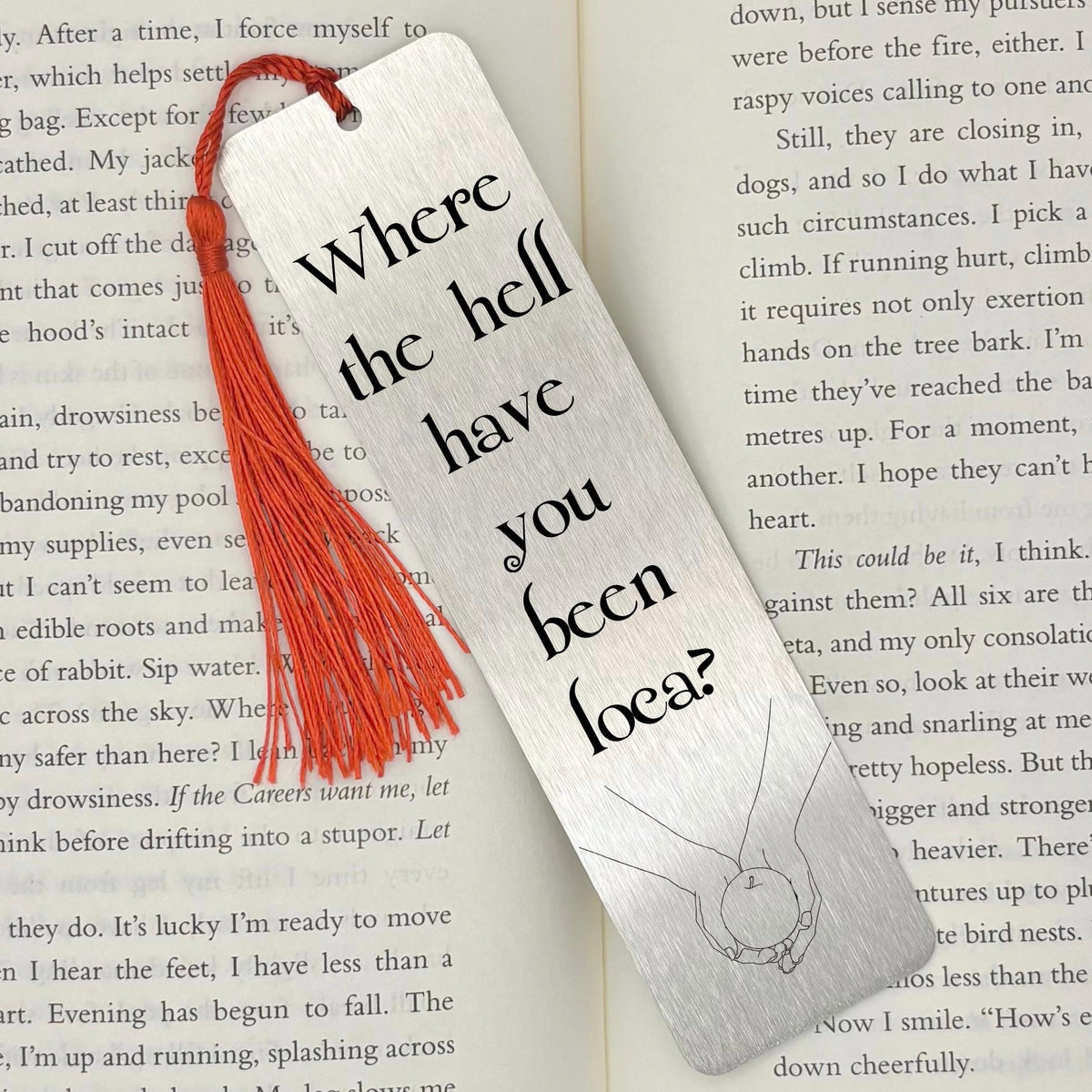     Where Have You Been Loca Metal Bookmark in brushed silver with black lettering and red tassel, displayed on an open book.