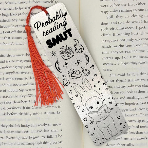 A brushed silver Probably Reading Smut Metal Bookmark on an open book with a red tassel