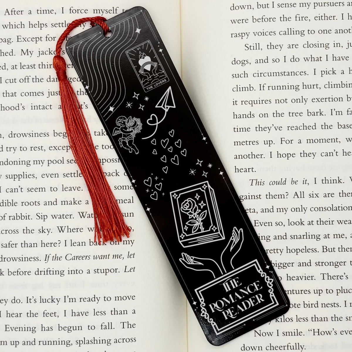 A black The Romance Reader Metal Bookmark on and open book with a red tassel