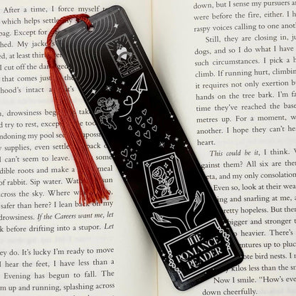 A black The Romance Reader Metal Bookmark on and open book with a red tassel