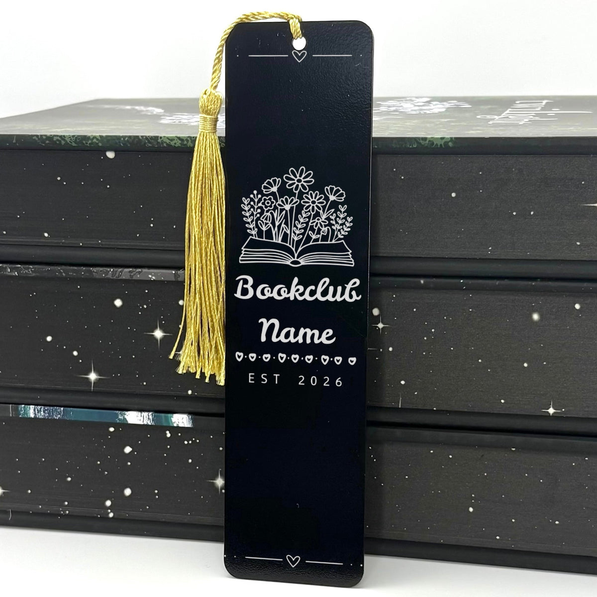 A black Personalised Book Club Name Metal Bookmark with gold tassel standing against stack of books