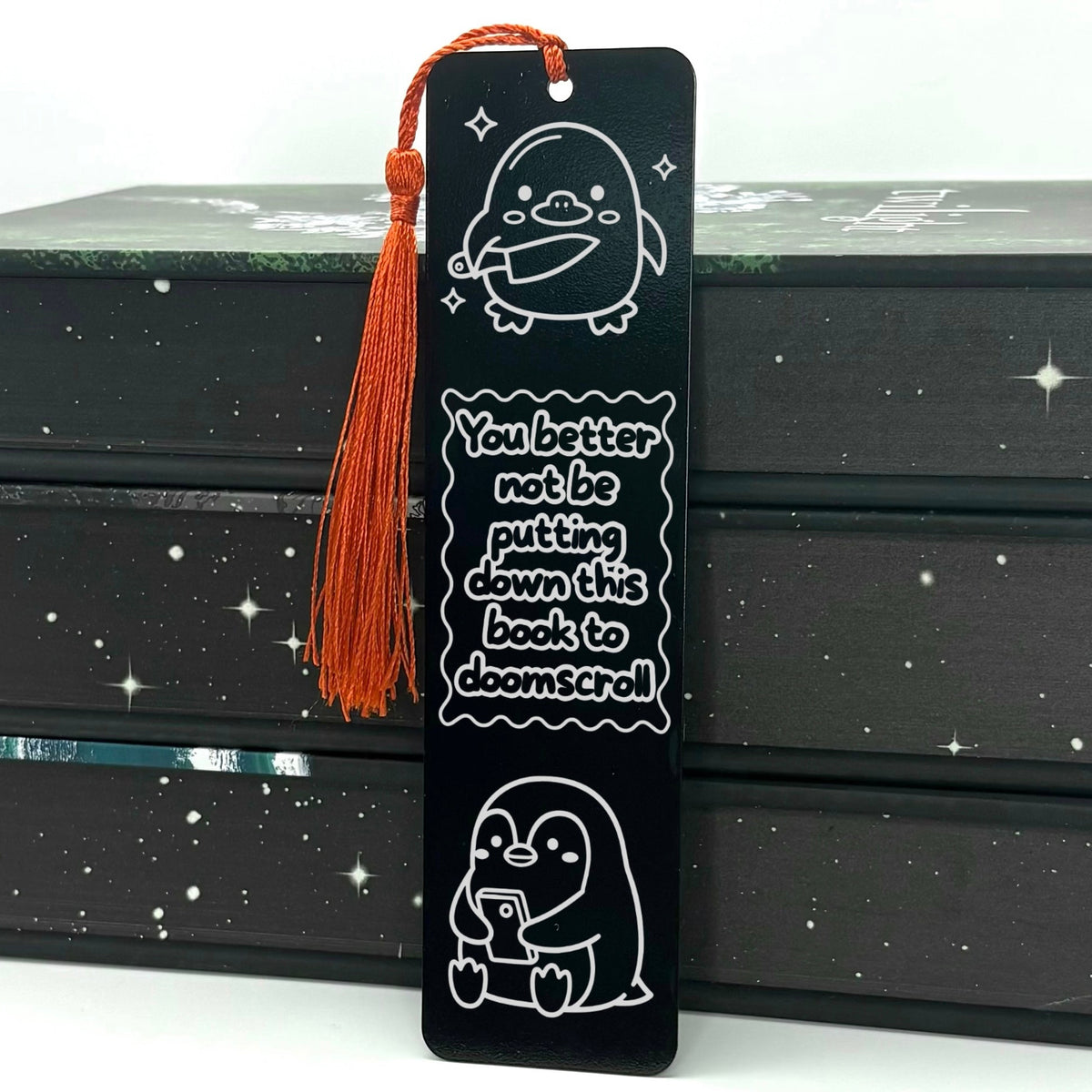 A black Doomscrolling Duck Metal Bookmark with red tassel standing against stack of bookw