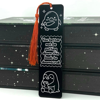 A black Doomscrolling Duck Metal Bookmark with red tassel standing against stack of bookw