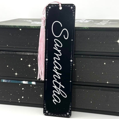 A black Personalised Name Metal Bookmark with pink tassel standing against a stack of books