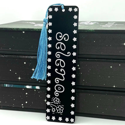 Black Personalised Name Floral Metal Bookmark with light blue tassel standing against stack of books