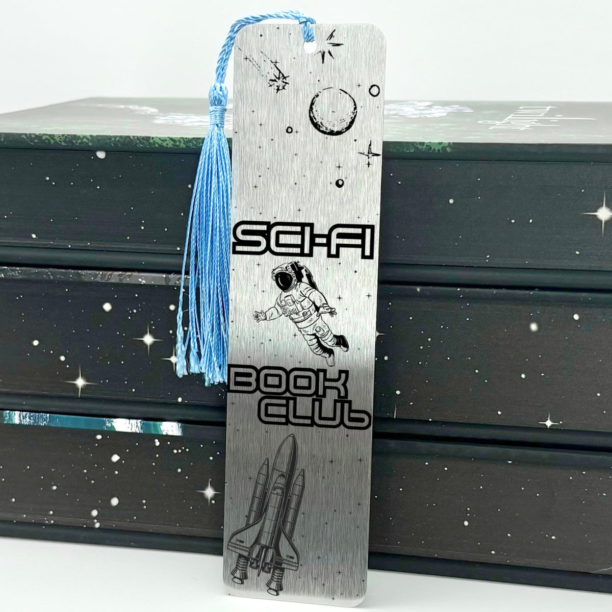     Sci-Fi Book Club Metal Bookmark in silver featuring engraved astronaut, moon, stars and space shuttle with blue tassel.