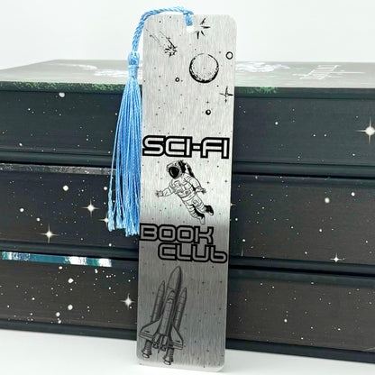     Sci-Fi Book Club Metal Bookmark in silver featuring engraved astronaut, moon, stars and space shuttle with blue tassel.