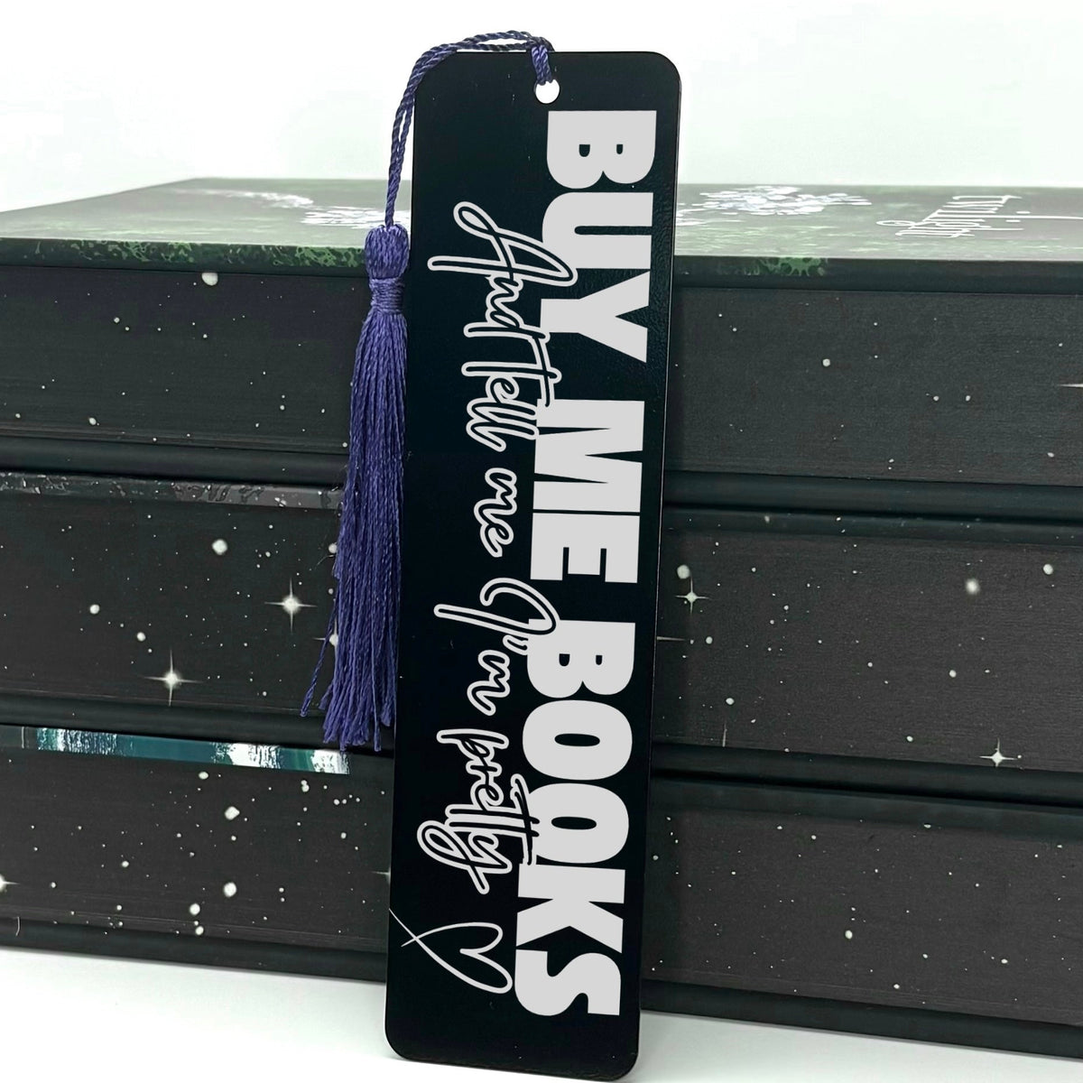 A black Buy Me Books And Tell Me I’m Pretty Metal Bookmark with purple tassel standing against stack of books