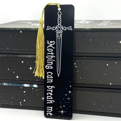 A black Nothing Can Break Me Sword Metal Bookmark with gold tassel standing against a backdrop of books
