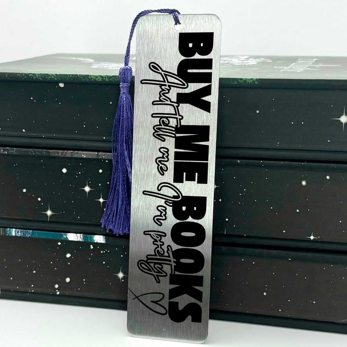 A brushed silver Buy Me Books And Tell Me I’m Pretty Metal Bookmark with purple tassel standing against stack of books