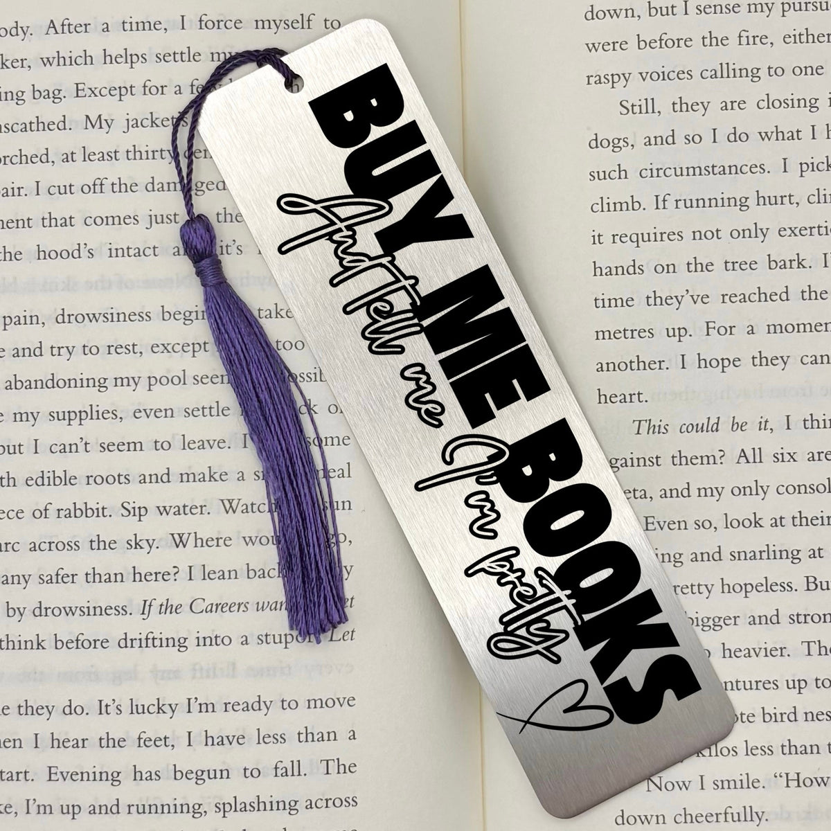 A brushed silver Buy Me Books And Tell Me I’m Pretty Metal Bookmark Metal Bookmark with purple tassel on an open book
