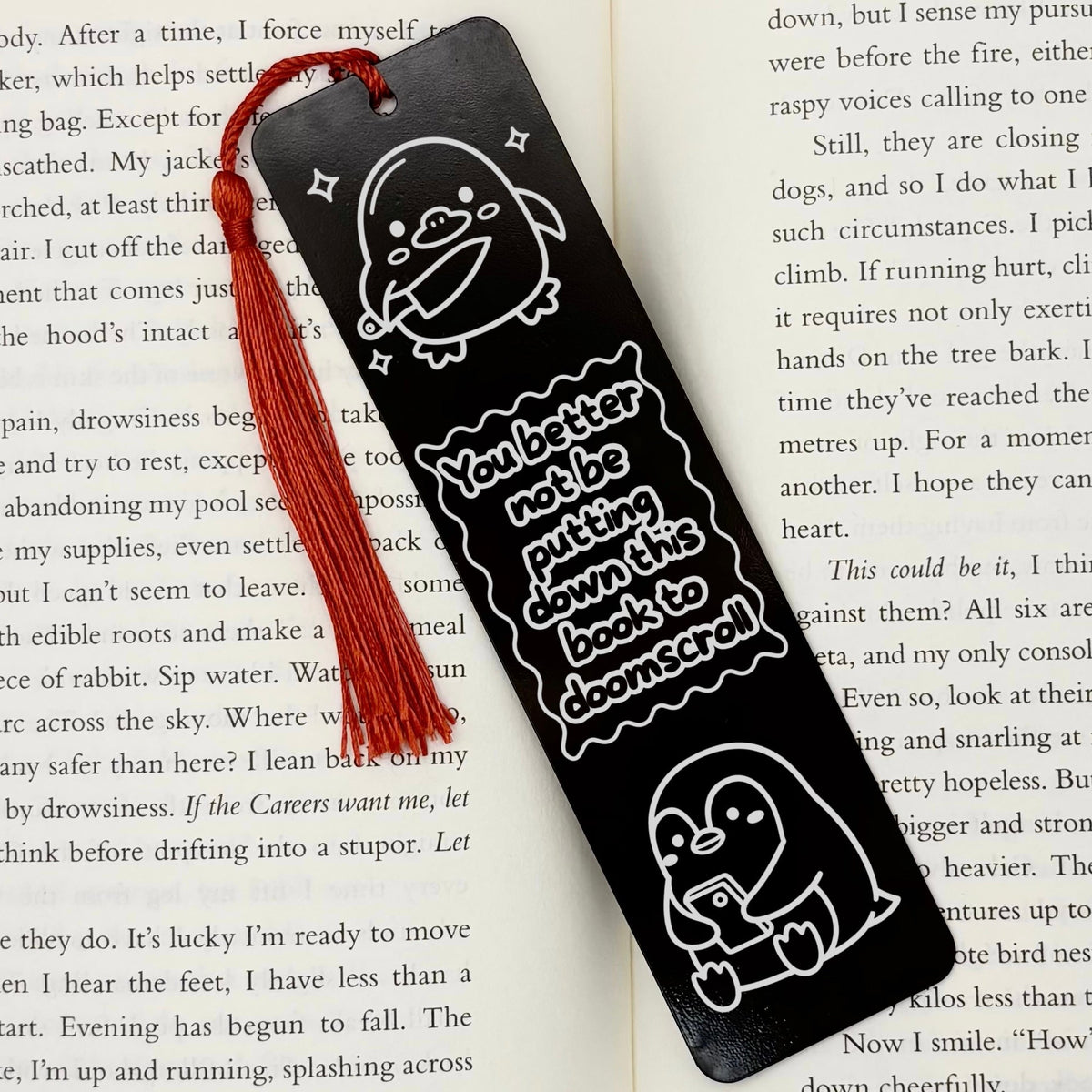 A black Doomscrolling Duck Metal Bookmark with red tassel on open book