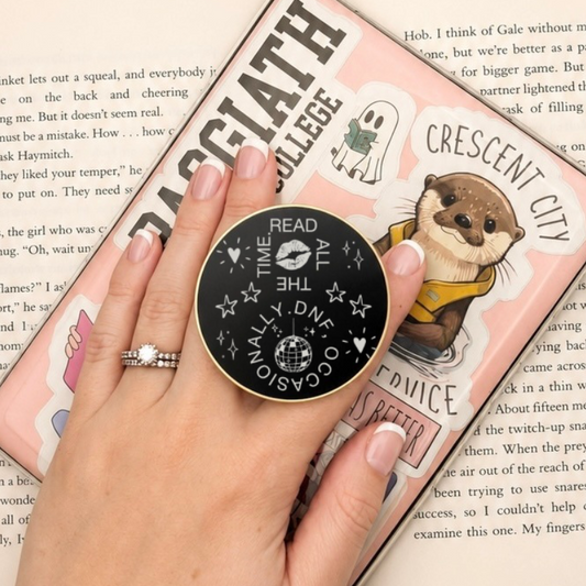     Read All The Time DNF Occasionally Metal MagSafe Kindle Grip held on a decorated Kindle while reading an open book.