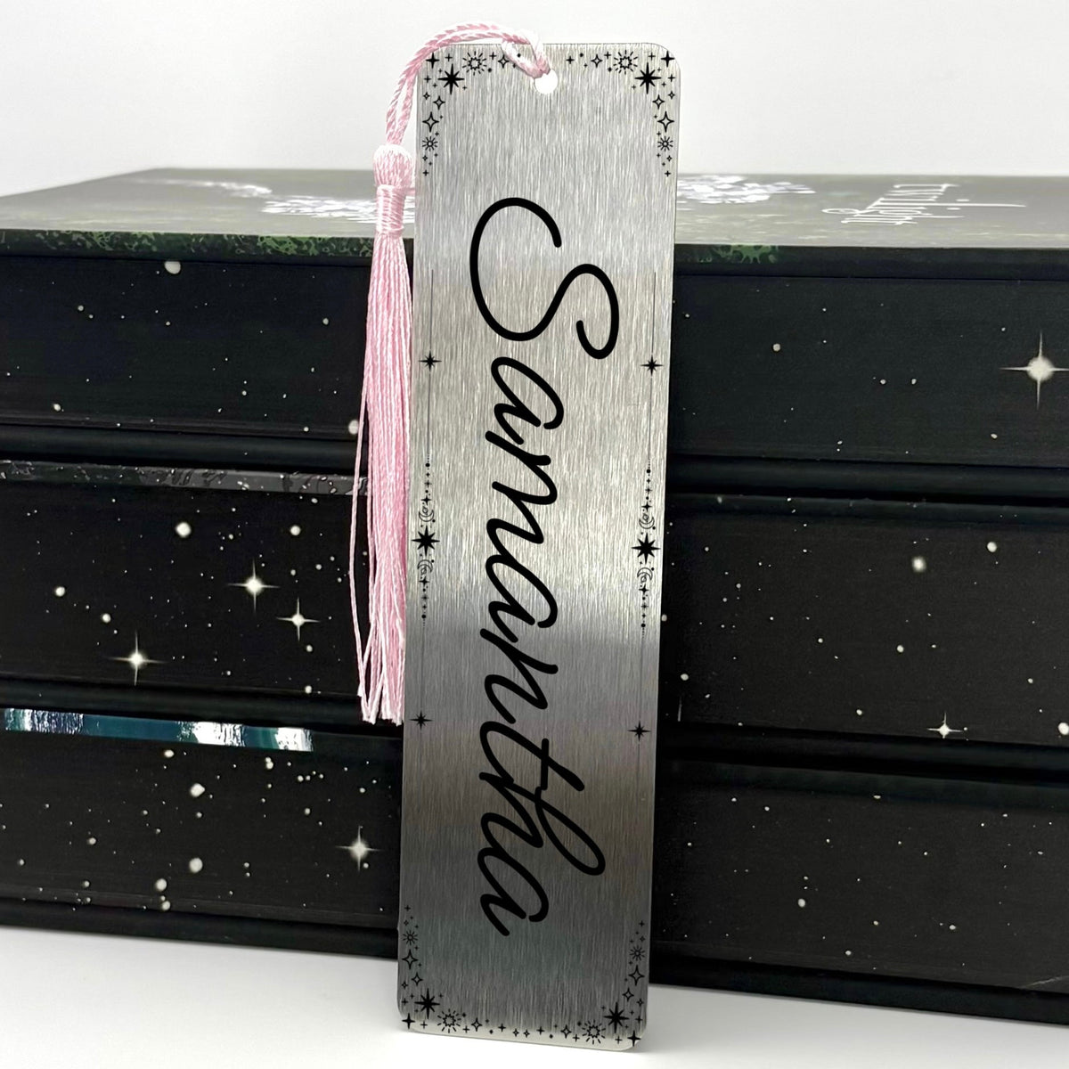 A brushed silver Personalised Name Metal Bookmark with pink tassel standing against a backdrop of books