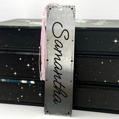 A brushed silver Personalised Name Metal Bookmark with pink tassel standing against a backdrop of books
