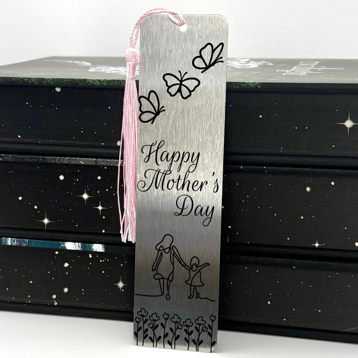 A brushed silver Happy Mothers Day Metal Bookmark with pink tassel standing against a stack of books