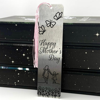 A brushed silver Happy Mothers Day Metal Bookmark with pink tassel standing against a stack of books