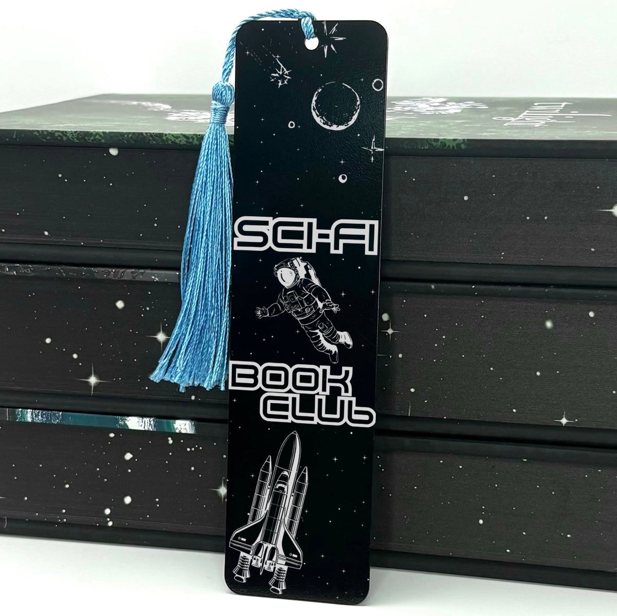     Sci-Fi Book Club Metal Bookmark in black with astronaut, moon, stars and space shuttle design with blue tassel.