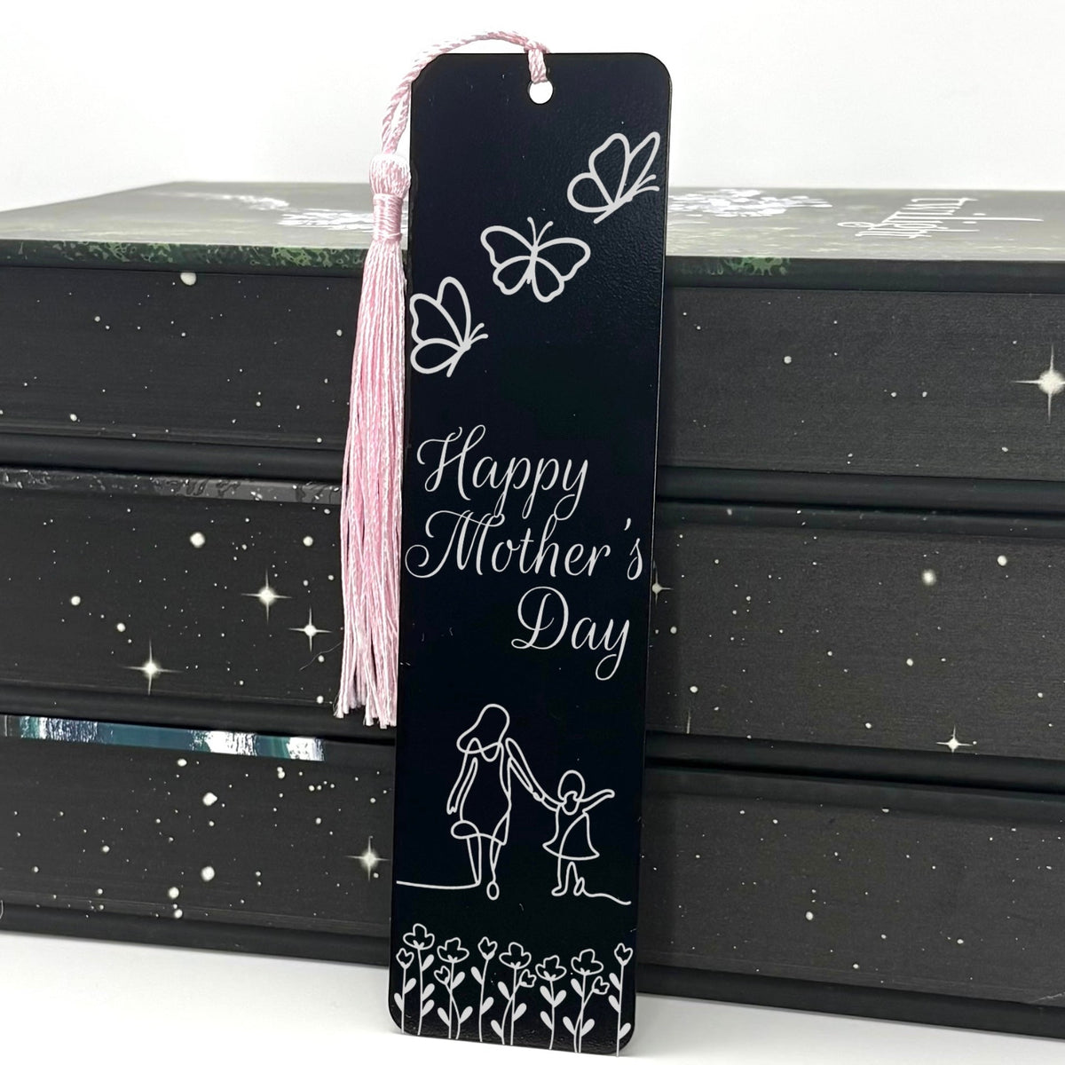 A black Happy Mothers Day Metal Bookmark with pink tassel standing against a stack of books