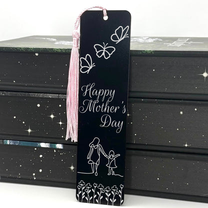 A black Happy Mothers Day Metal Bookmark with pink tassel standing against a stack of books