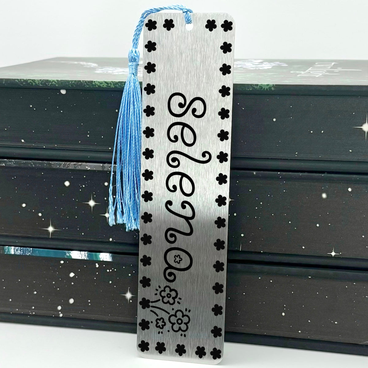Brushed silver Personalised Name Floral Metal Bookmark with light blue tassel standing against stack of books