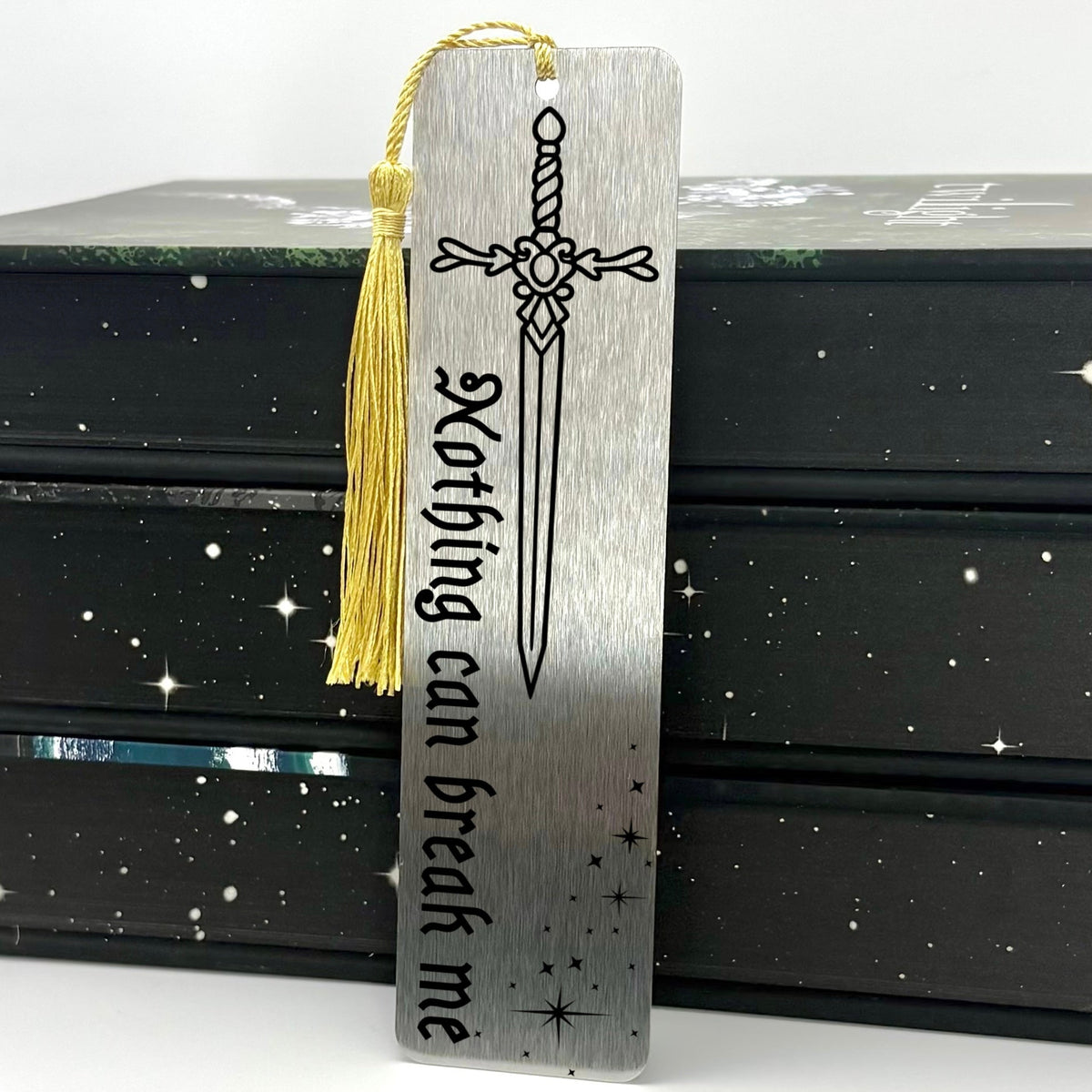 A brushed silver Nothing Can Break Me Sword Metal Bookmark with gold tassel standing against a backdrop of books