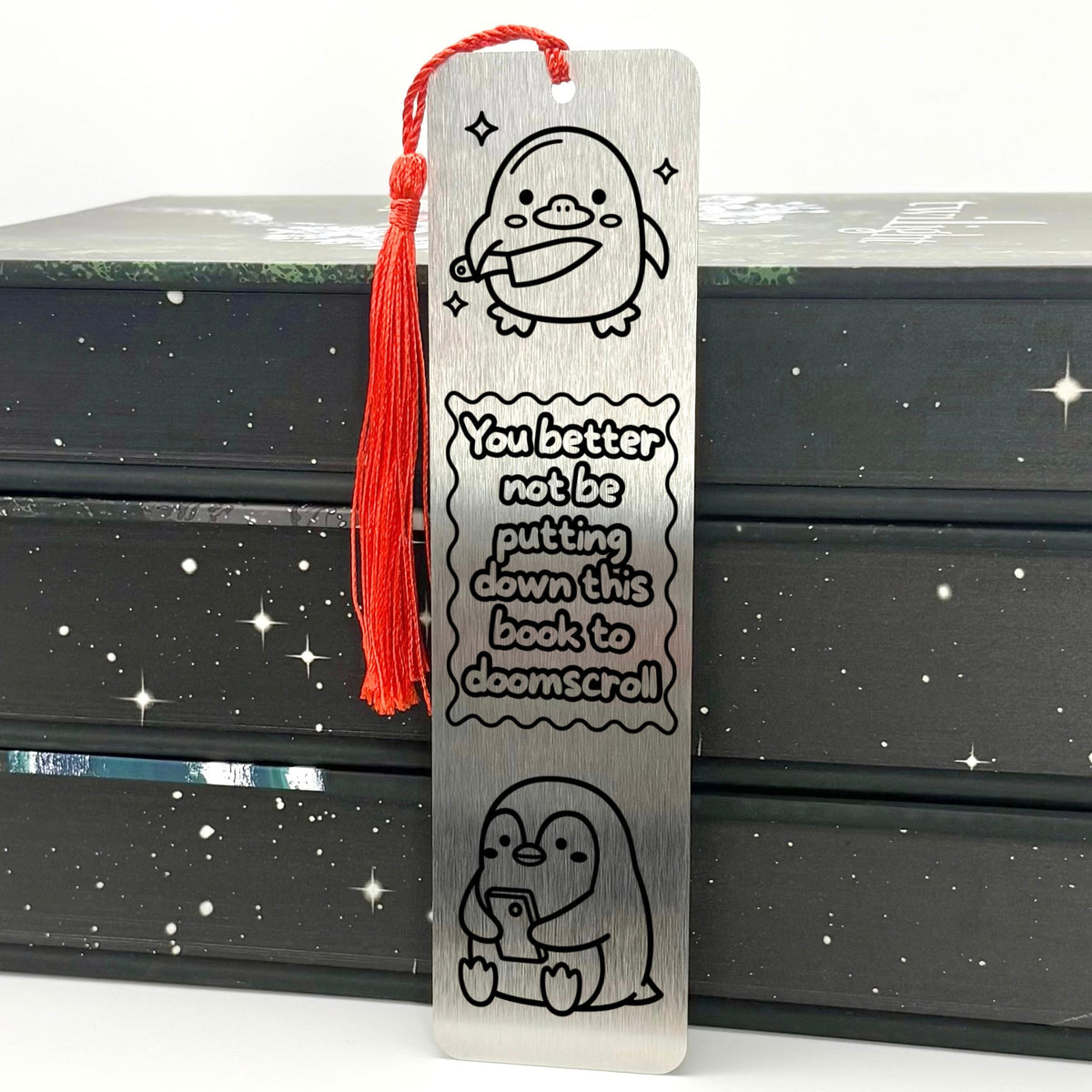 A brushed silver Doomscrolling Duck Metal Bookmark with red tassel standing against stack of books