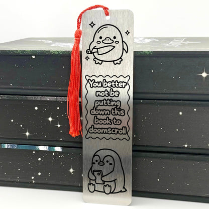 A brushed silver Doomscrolling Duck Metal Bookmark with red tassel standing against stack of books