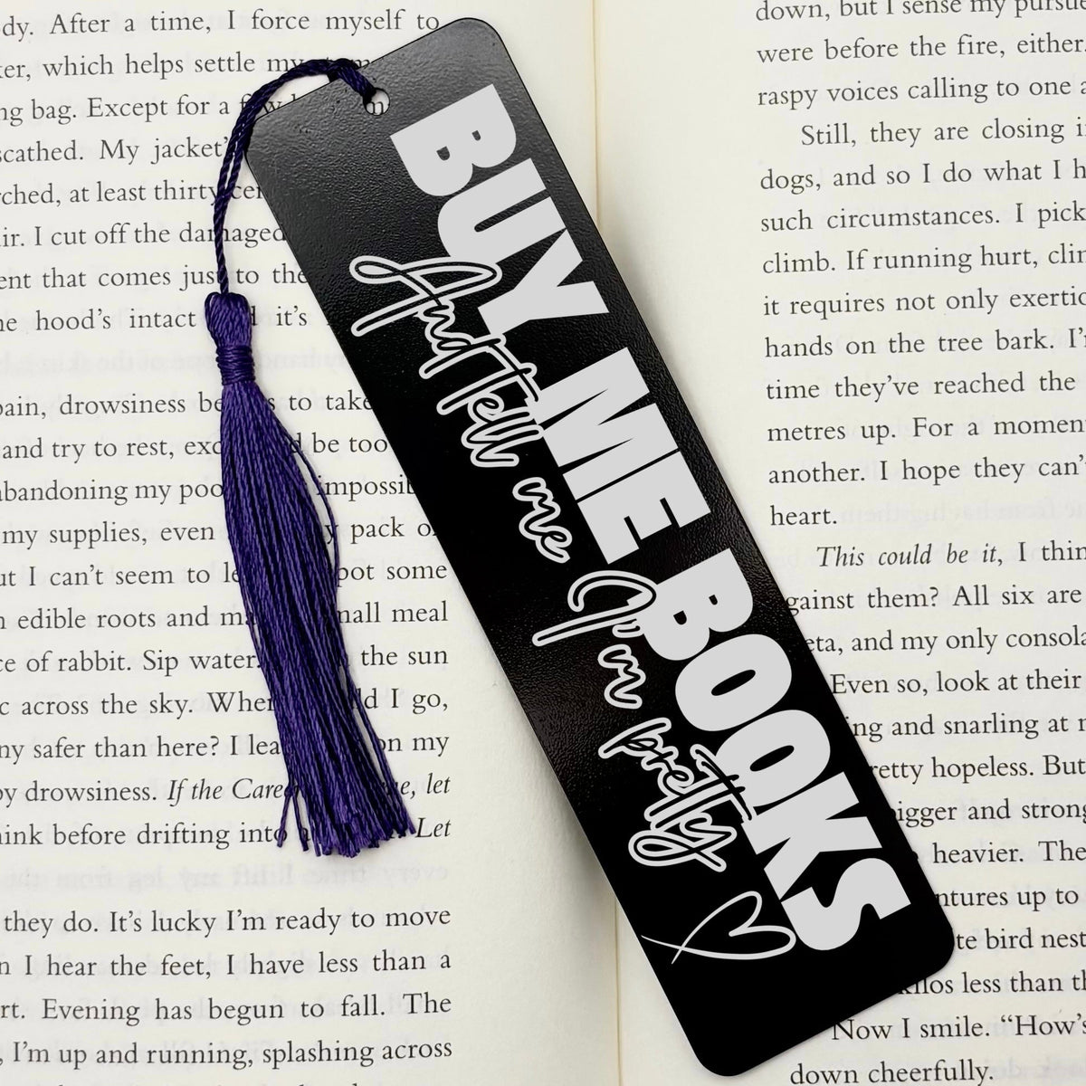 A black Buy Me Books And Tell Me I’m Pretty Metal Bookmark with purple tassel on an open book