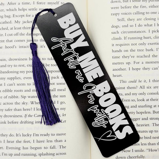A black Buy Me Books And Tell Me I’m Pretty Metal Bookmark with purple tassel on an open book