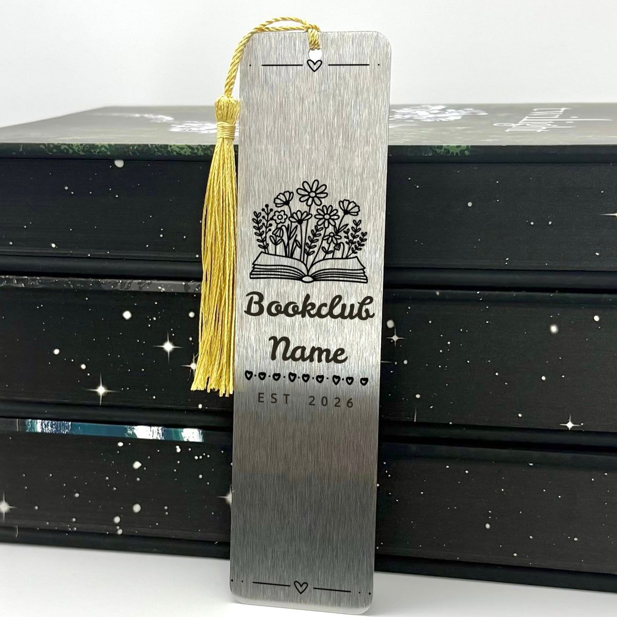 A brushed silver Personalised Book Club Name Metal Bookmark with gold tassel standing against stack of books
