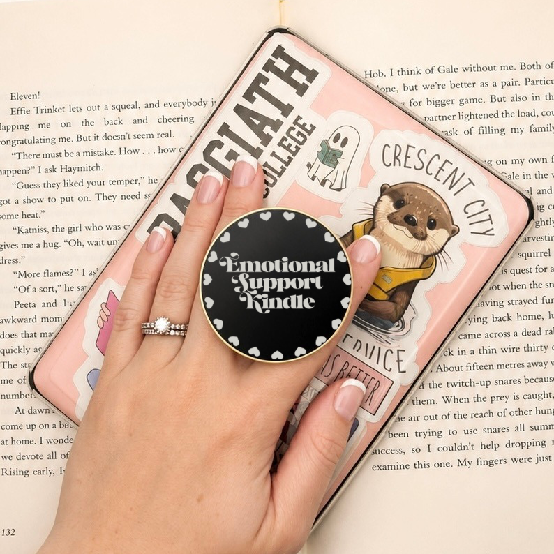 Hand holding an Emotional Support Kindle Metal E-Reader Grip on a Kindle, featuring colorful stickers and a cozy reading atmosphere.
