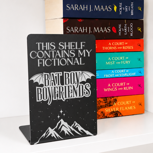 Bat Boys Metal Bookshelf Sign Bookend with engraved bat wings, stars and mountain design displayed in front of a colourful stack of fantasy books.