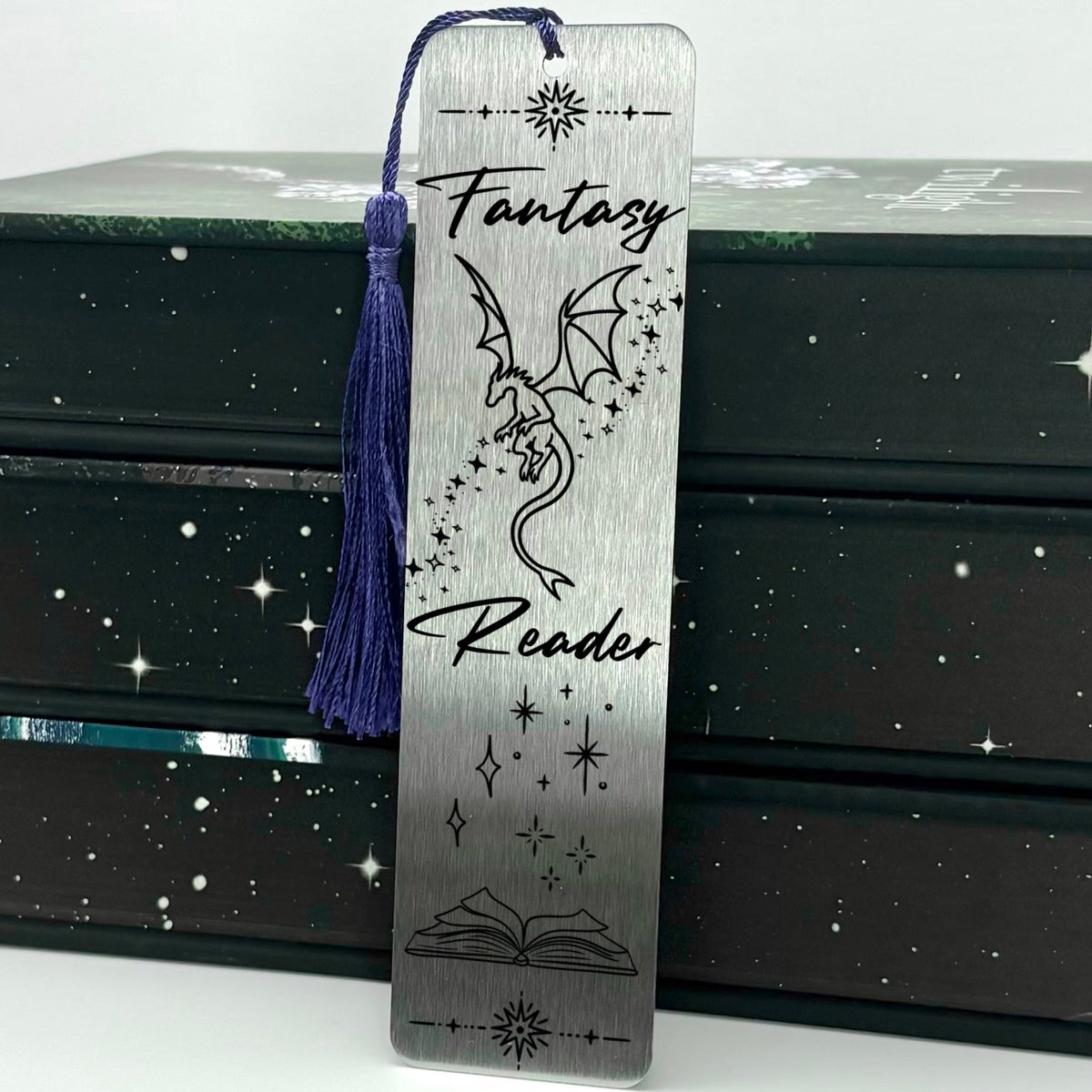 A brushed silver Fantasy Reader Dragon Metal Bookmark with purple tassel standing against a backdrop of books