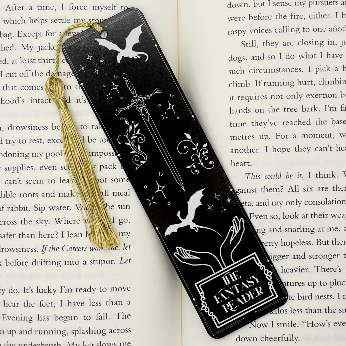 A black The Fantasy Reader Metal Bookmark on and open book with a gold tassel
