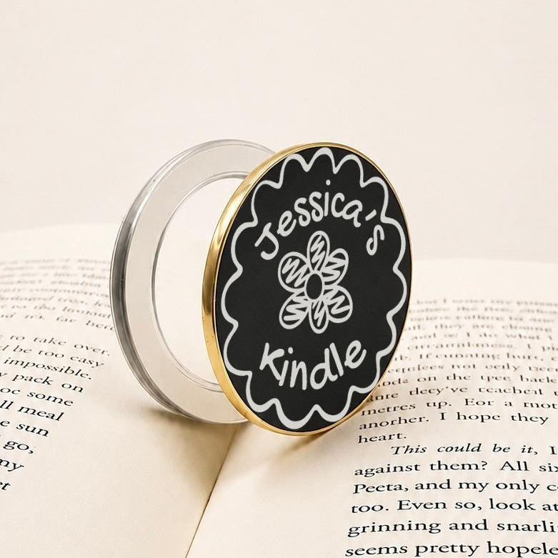 Personalised Name Floral Metal E-Reader Grip featuring a black and gold design for Jessica's Kindle on an open book.