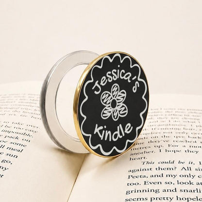 Personalised Name Floral Metal E-Reader Grip featuring a black and gold design for Jessica's Kindle on an open book.